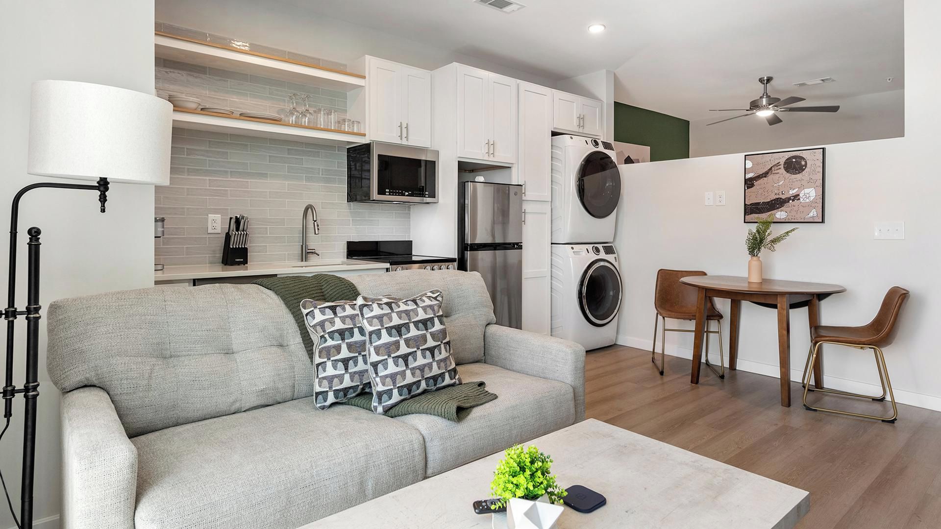A modern apartment with a grey sofa, a coffee table, a dining area with a table and chairs, a kitchen with appliances, and a ceiling fan.