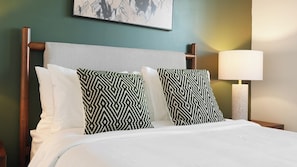 Quarto de hotel com cama, duas almofadas estampadas, mesa de cabeceira, abajur e pintura na parede.
