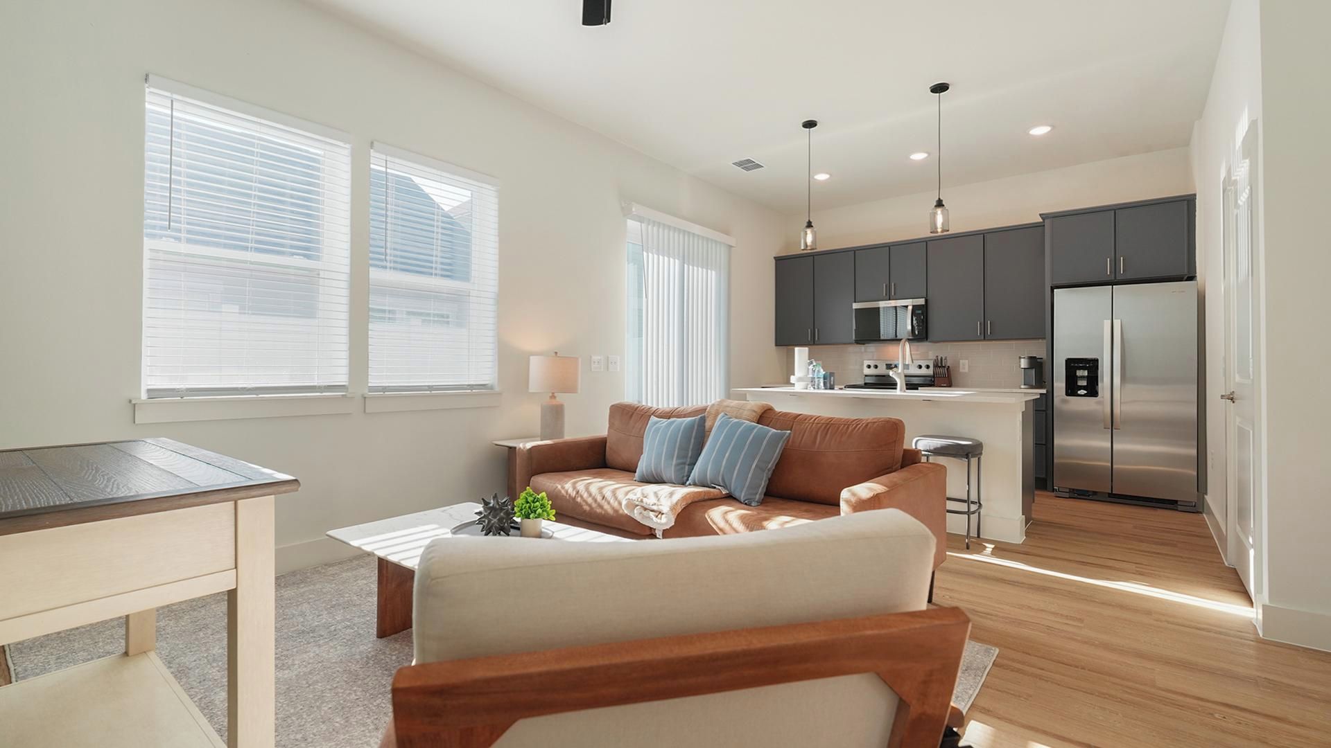 A modern living room with a sofa, coffee table, and a dining table. There is a kitchen area with a refrigerator, microwave, and sink.