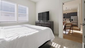 A bedroom with a bed, a dresser, a television, and a view into a kitchen area. - Landing | Stunning 2BD, Gym (Palmetto)