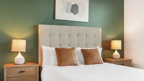 Een net opgemaakt bed met een geborduurde hoofdbord, twee nachtkastjes met lampen, en een abstract kunstwerk aan de muur.