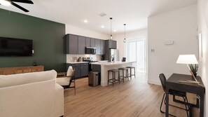 A modern kitchen with dark cabinets, a central island with stools, a dining table with chairs, and a flat-screen TV mounted on the wall. - Landing | Chic 2BD, Gym (Palmetto)