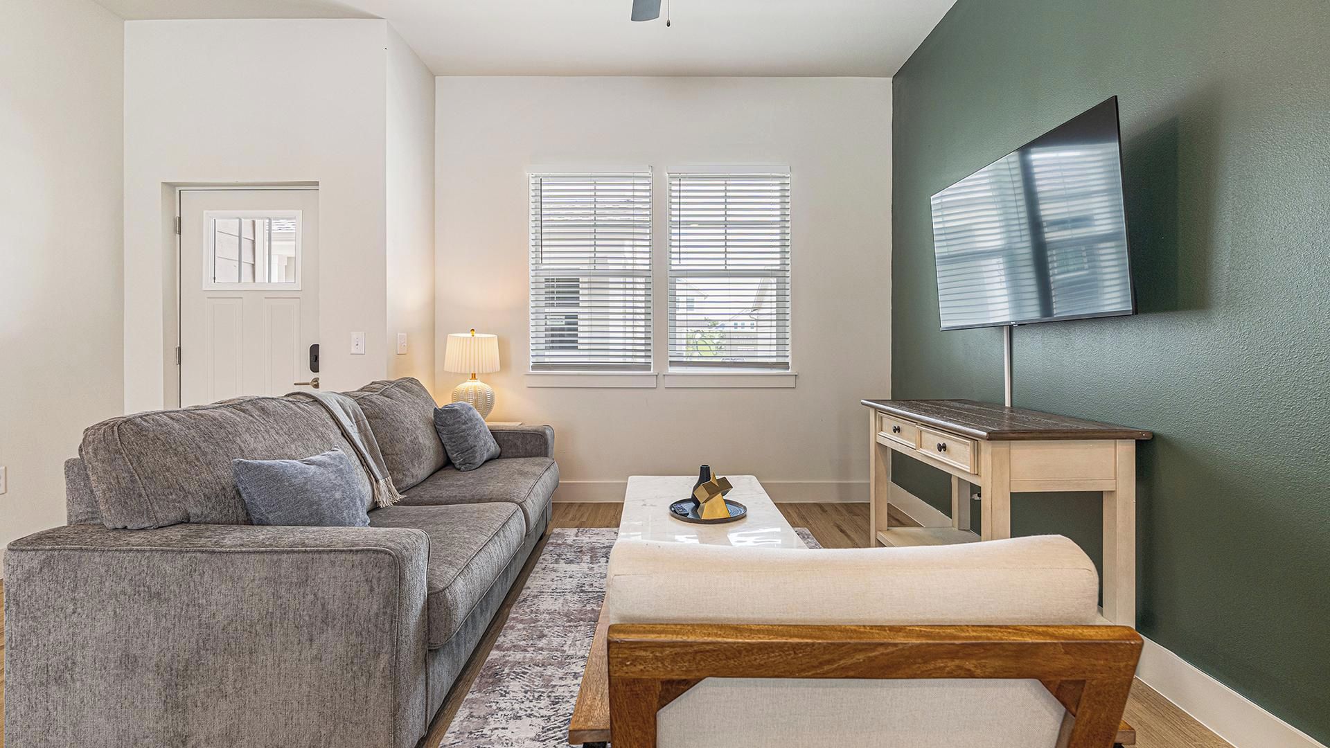 Ein Wohnzimmer mit grauem Sofa, einem Couchtisch, einer Lampe und einem an der Wand befestigten Fernseher.