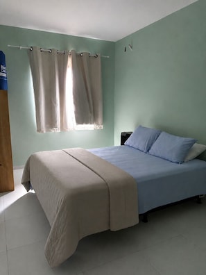 Een slaapkamer met een bed, nachtkastje, raam met gordijnen en een houten kledingkast.