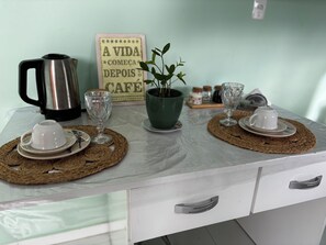 A kitchen counter with a coffee maker, two cups, a potted plant, and a sign. - Praia do Francês: sun, sea, the lightness of being where the soul finds its balance. (Marechal Deodoro)