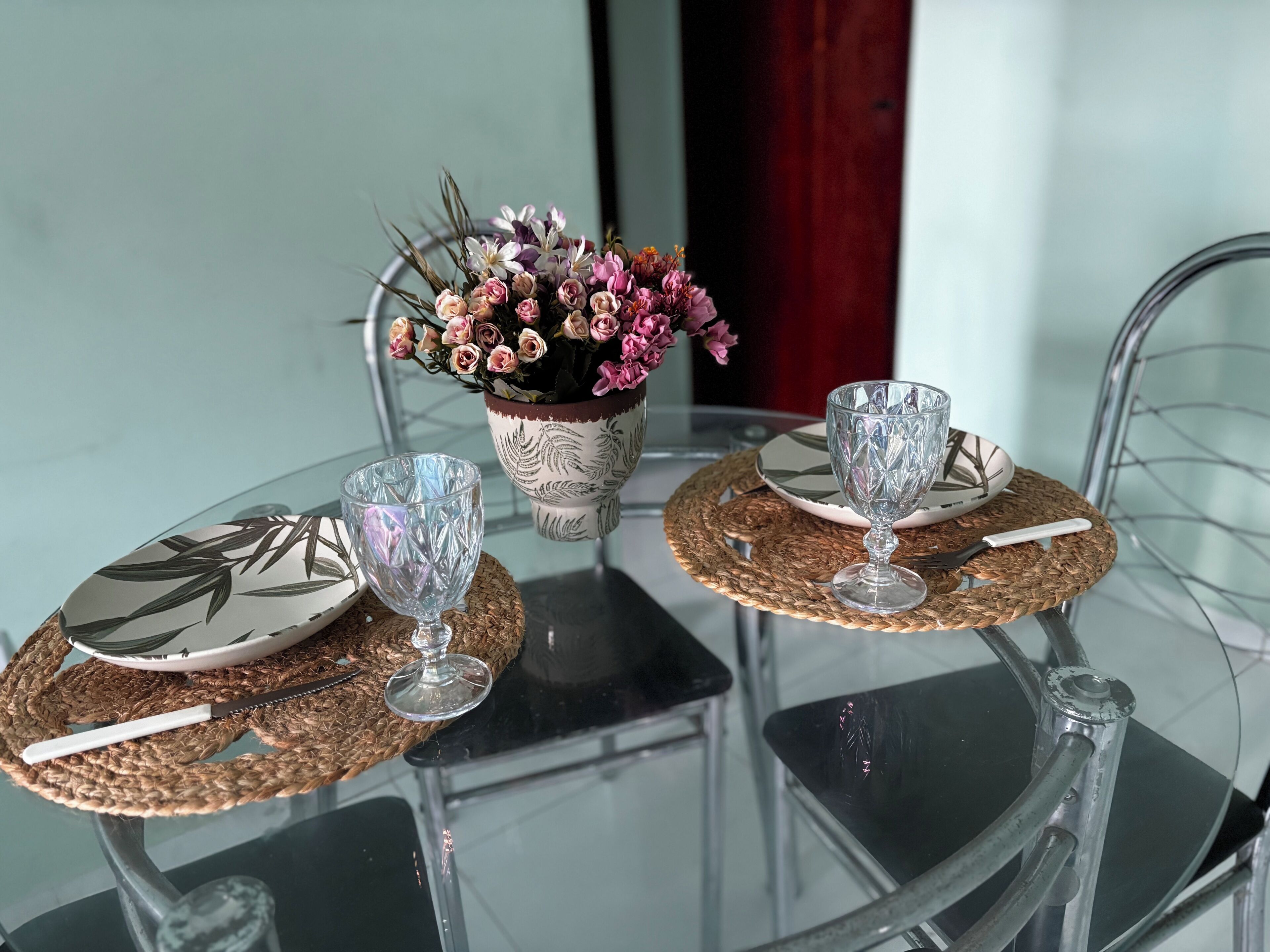 A glass table with two plates, two glasses, and a decorative vase with flowers.