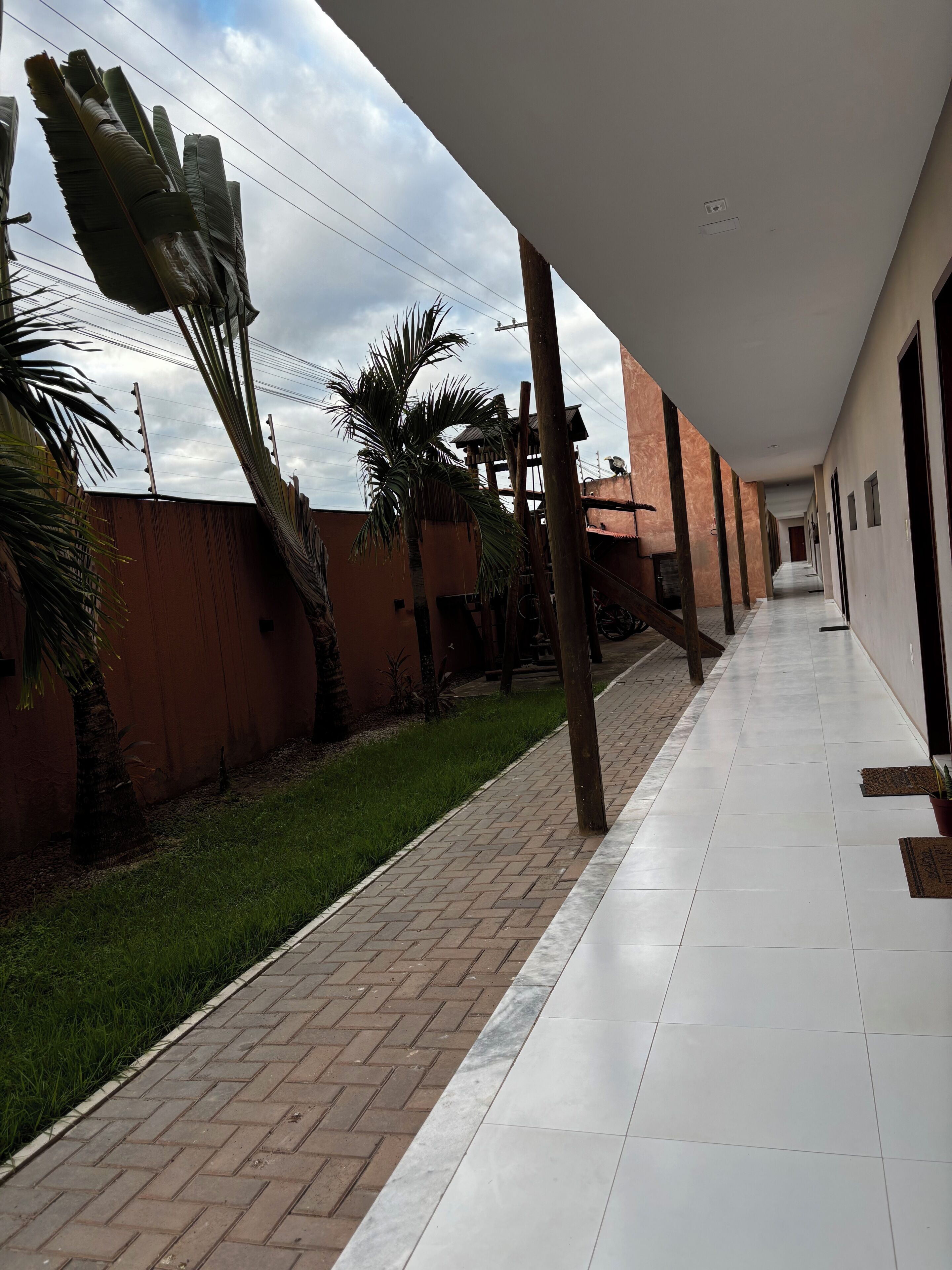 Um corredor coberto com piso de azulejos, caminho de tijolos e uma palmeira.