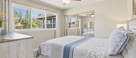 A bedroom with a bed, a desk, a ceiling fan, a window, and a mirror.