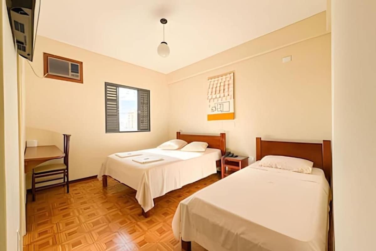 A hotel room with two beds, a desk, a chair, a window with blinds, and a wall-mounted artwork.