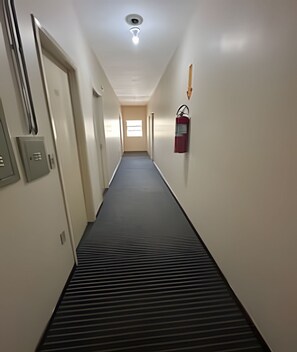 Un pasillo estrecho con un extintor montado en la pared, un respiradero en el suelo y una puerta a la izquierda.