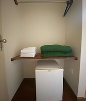 Una habitación pequeña con un refrigerador blanco, un estante de madera con toallas blancas y verdes dobladas, y una puerta con manija.