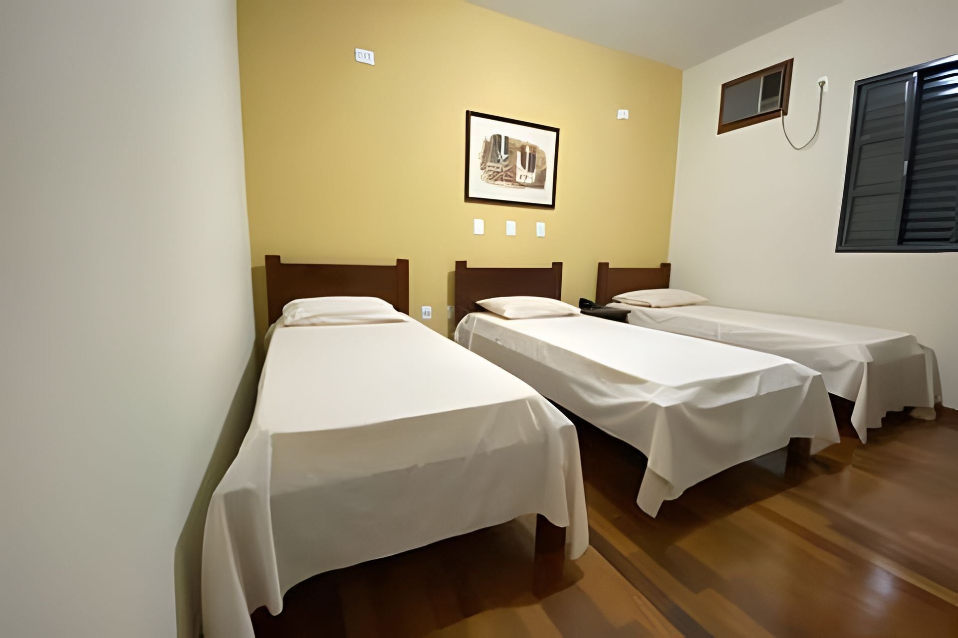 Quarto com três camas de solteiro, um quadro pendurado na parede e uma janela com persianas.