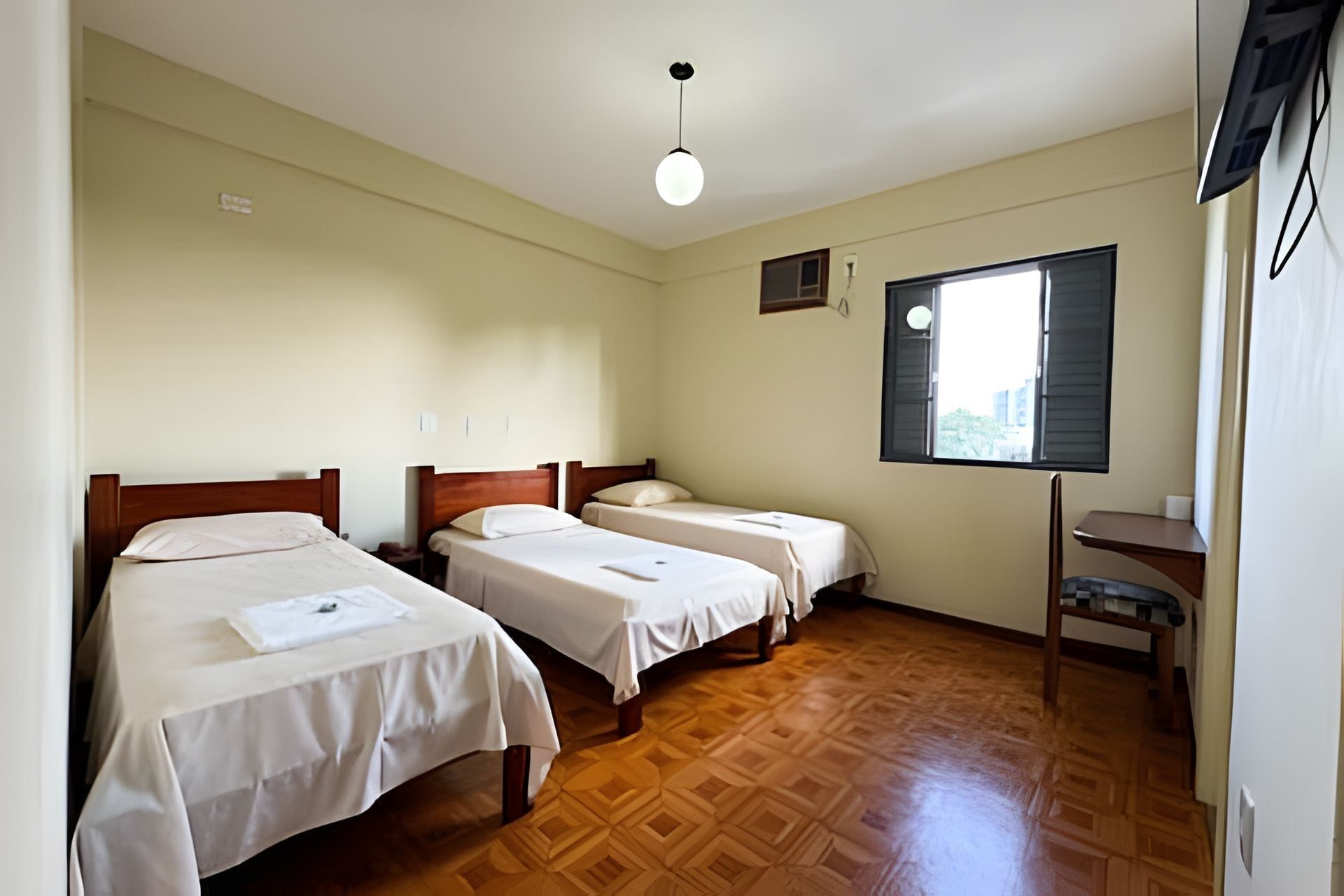 Una habitación con tres camas individuales, suelo de madera, ventana con persianas y un escritorio pequeño.
