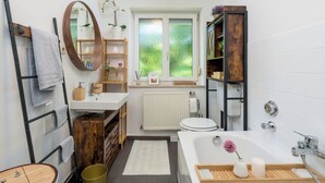 A modern bathroom with a wooden vanity, a freestanding bathtub, a toilet, and a window providing a view of greenery. - 1OG M schöne Wohnung 10 min von Salzburg (Freilassing)