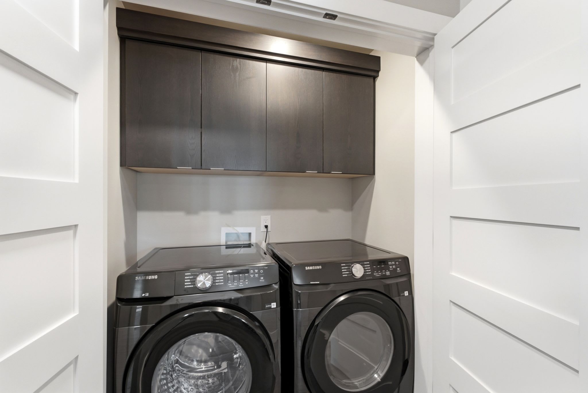Une buanderie équipée de deux machines à laver Samsung, avec un meuble en bois foncé au-dessus et une porte blanche sur le côté gauche.
