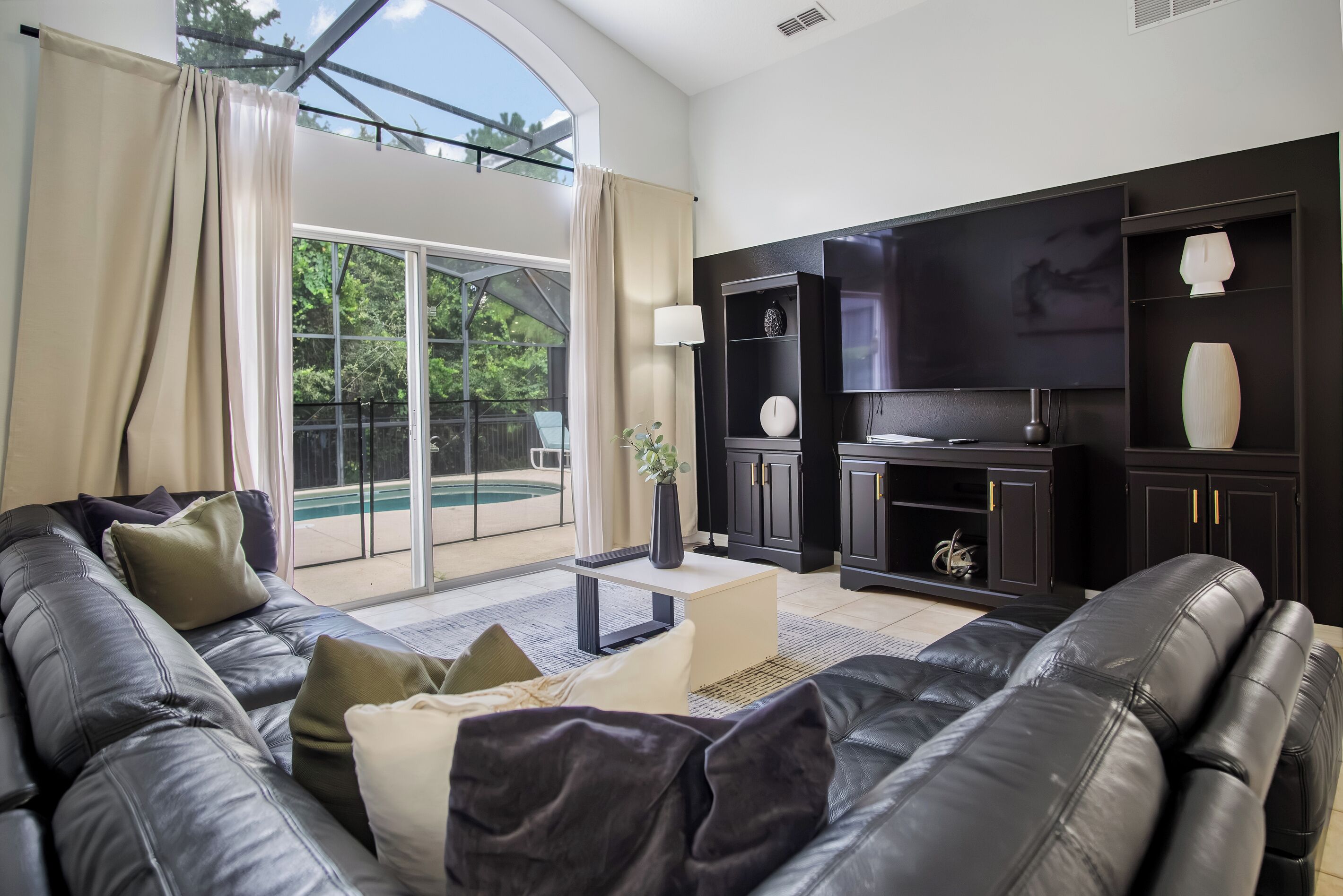 Een moderne woonkamer met een zwarte leren bank, een flatscreen televisie en een zwembad zichtbaar door de glazen deuren.