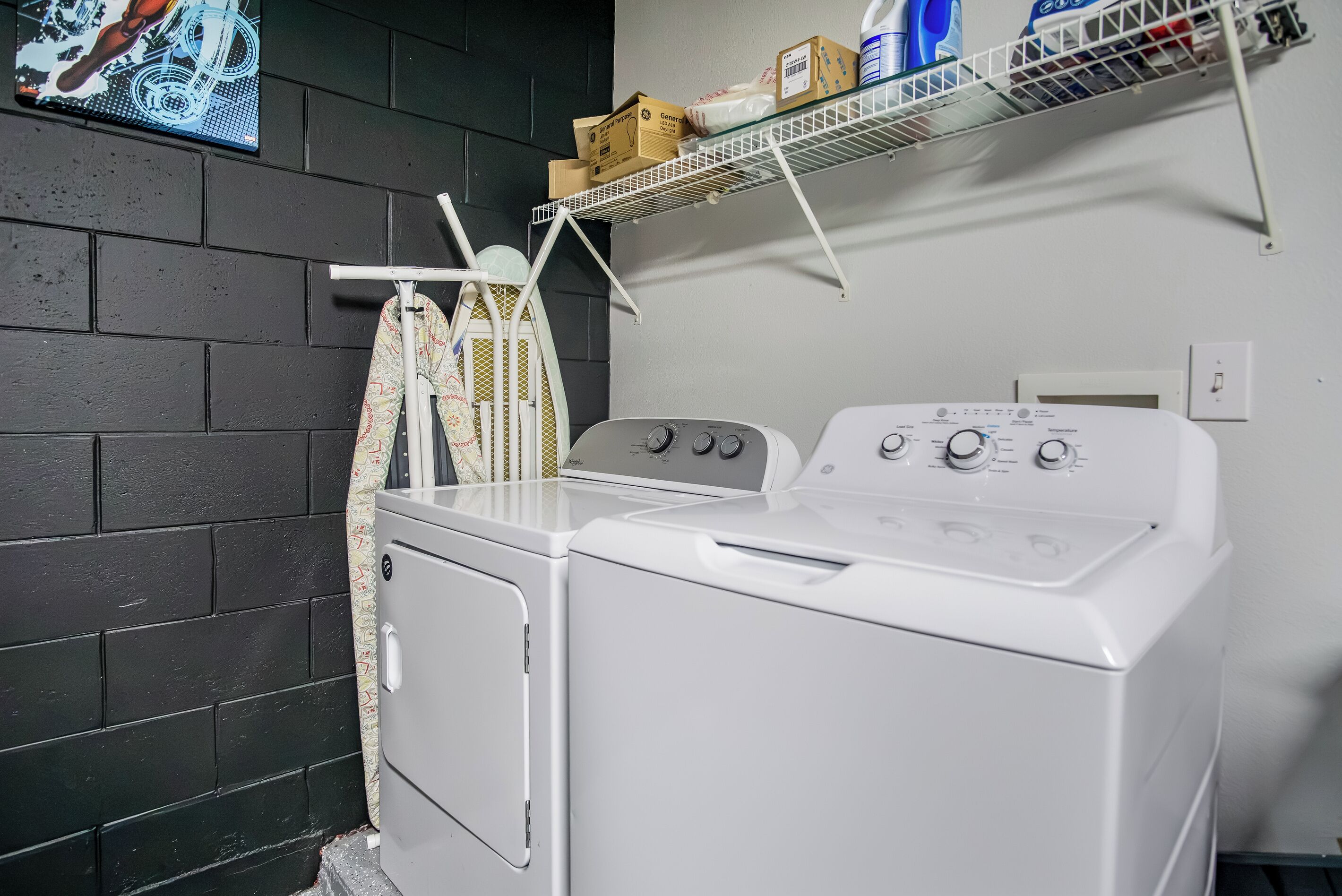 A laundry area with a white washing machine and dryer, a drying rack with clothes, and a shelf with boxes and cleaning supplies.