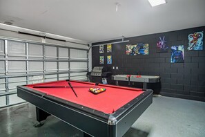 A pool table with a red felt surface, a cue stick, and a ball rack in a room with a black brick wall and comic book art. - Luxury 5BD home close to Disney with Private Pool (Kissimmee)