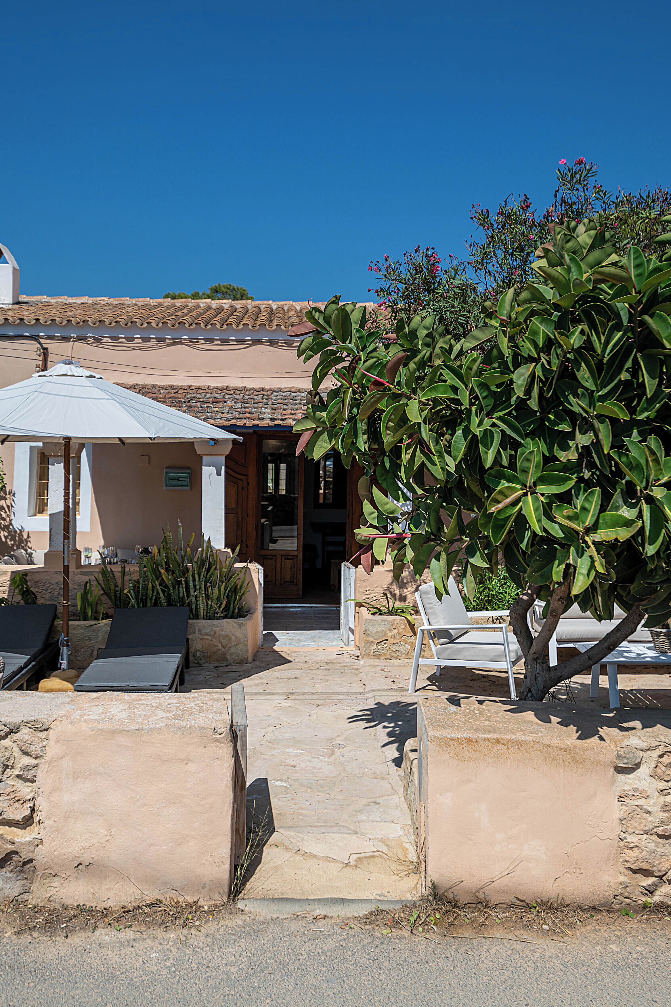 A building with a tiled roof, outdoor seating with chairs and tables, and a large tree in the foreground.