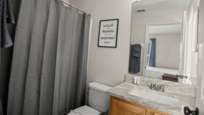 Une salle de bain avec un rideau de douche, des toilettes, un lavabo avec un miroir et une citation encadrée au mur.