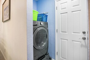 Un espace dédié à la lessive, comprenant un lave-linge à chargement frontal, un panier bleu et un seau vert.