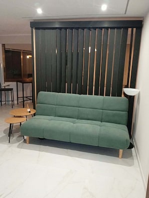 A green sofa in a room with a wooden slatted partition, a small round table, and a floor lamp. - Vrbo Property (Castelldefels)