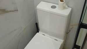 Un bagno con rotolo di carta igienica sopra, parete a effetto marmo e porta doccia in vetro.