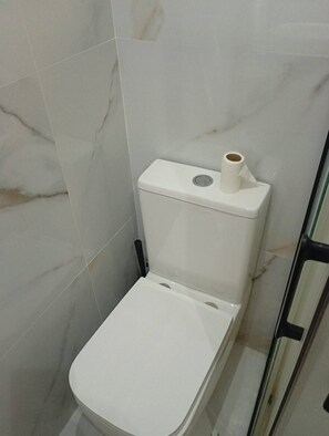A toilet with a roll of toilet paper on top, a marble-patterned wall, and a glass shower door. - Vrbo Property (Castelldefels)