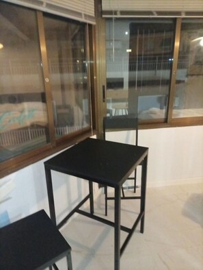 A room with a table and two chairs, a window with blinds, and a view of a building outside. - Vrbo Property (Castelldefels)