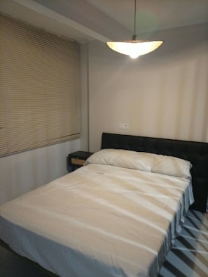 A bedroom with a bed, a nightstand, a lamp, and a window with blinds. - Vrbo Property (Castelldefels)
