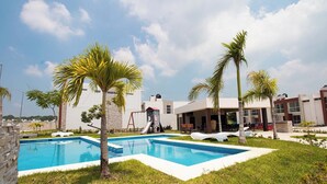 A swimming pool with a white deck, surrounded by grass and palm trees, with a building in the background. - Santa Ana el Dorado (San José Novillero)