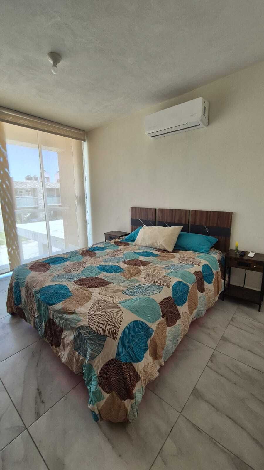 A bedroom with a bed, a nightstand, an air conditioning unit, and a view through a sliding door.