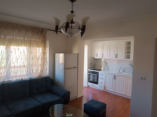 A modern living room with a sofa and a glass coffee table, a kitchen with white cabinets and appliances, and a ceiling fan.