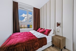 Ein Schlafzimmer mit Bett, Nachttisch, einem Fenster mit Vorhängen und Blick auf Himmel und Wolken.