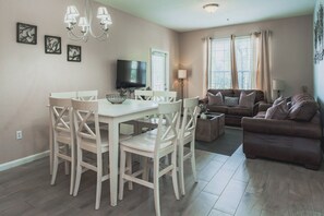 Un salon avec un canapé marron, un téléviseur et un coin repas comprenant une table et des chaises blanches.