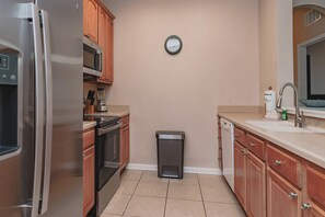 A compact kitchen with a stainless steel refrigerator, microwave, oven, and dishwasher. A clock is mounted on the wall, and a trash bin is placed against the wall. - Comfortable 3BR Vista Cay Near Universal (Orlando)