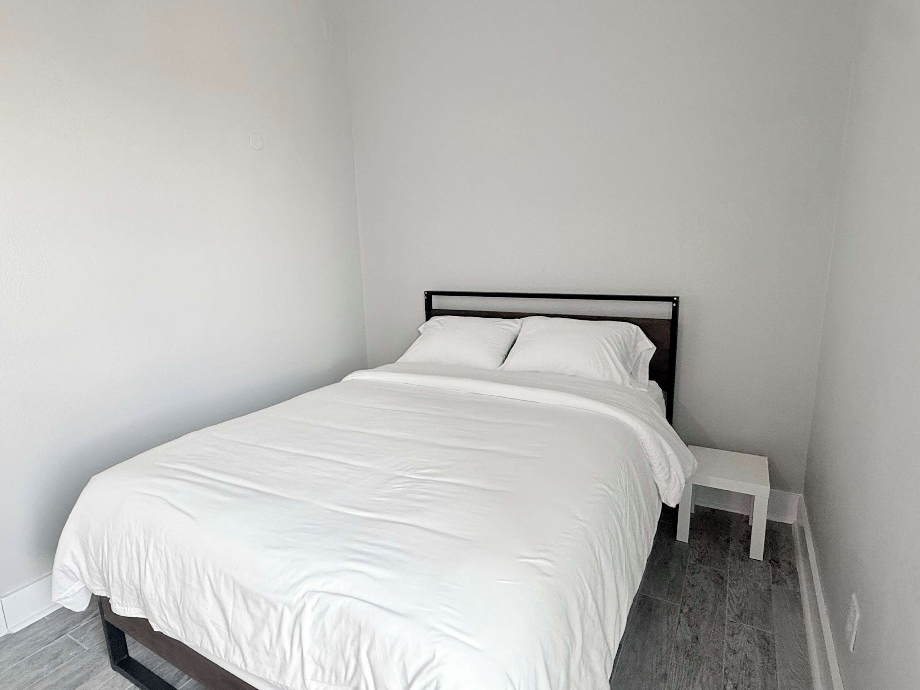 Un lit simple avec un matelas et des oreillers blancs, une structure en métal noir et une table de chevet blanche, dans une pièce aux murs blancs unis et au sol carrelé.