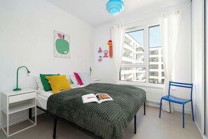 A bedroom with a bed, a chair, a nightstand, a window with a view of buildings, and a ceiling light. - Designer Apartment with Balcony | 1 Bedroom (Poznań)