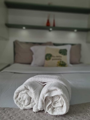 Ein ordentlich bezogenes Bett, im Vordergrund ein gefaltetes weißes Handtuch, ein Kissen mit grünem Muster und im Hintergrund eine Ablage mit zwei roten Flaschen.