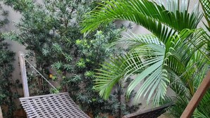 A hammock chair with a metal mesh seat and wooden frame, set against a backdrop of various green plants and a textured wall. - Studio Metrô Butantã (São Paulo)