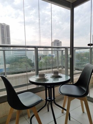 Ein Balkon mit Glastisch und zwei Stühlen, der auf eine Stadtlandschaft mit Gebäuden und Bäumen blickt.