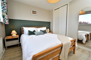Una cama bien hecha con sábanas blancas y almohadas verdes, una lámpara de noche y una mesita de madera.
