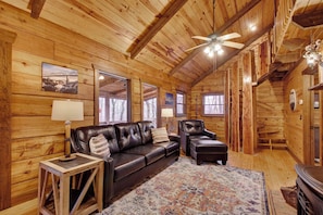 A cozy wooden cabin interior with a leather sofa, two armchairs, a rug, and a ceiling fan. - Log Cabin on 2 acres, hot tub, pond, fishing, pets, 1 mile TN Riverplace Venue (Chattanooga)
