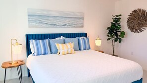 Une chambre à coucher avec un grand lit, une tête de lit, un tableau accroché au mur, une plante en pot et des lampes de chevet.