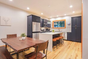 A modern kitchen with dark cabinets, stainless steel appliances, a wooden dining table, and a bar area with stools. - Modern Mt. Baker Cabin with Private Hot Tub + Fire Pit (Glacier)