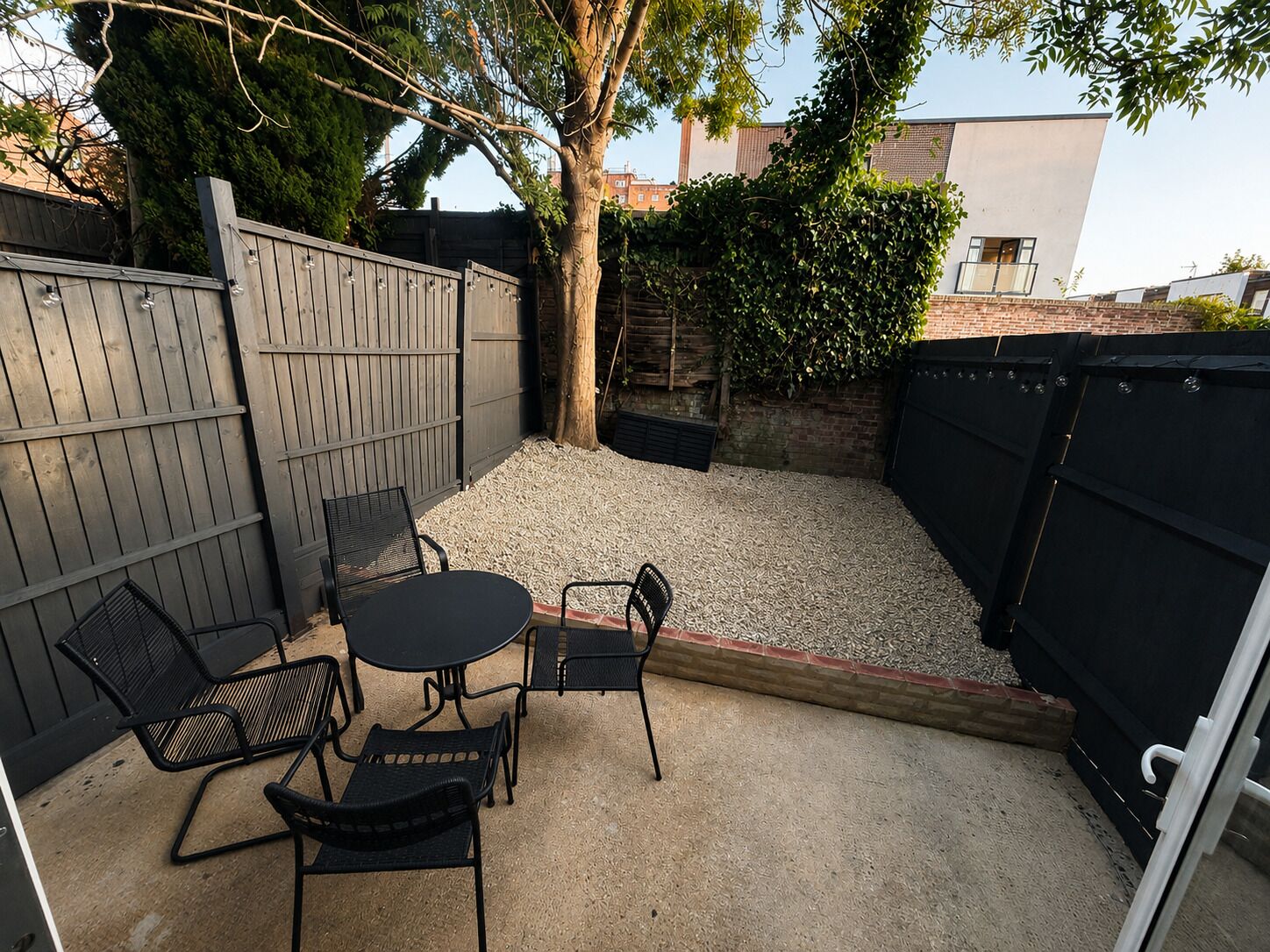 Un patio exterior con una mesa redonda y cuatro sillas, rodeado de grava y una cerca de madera.