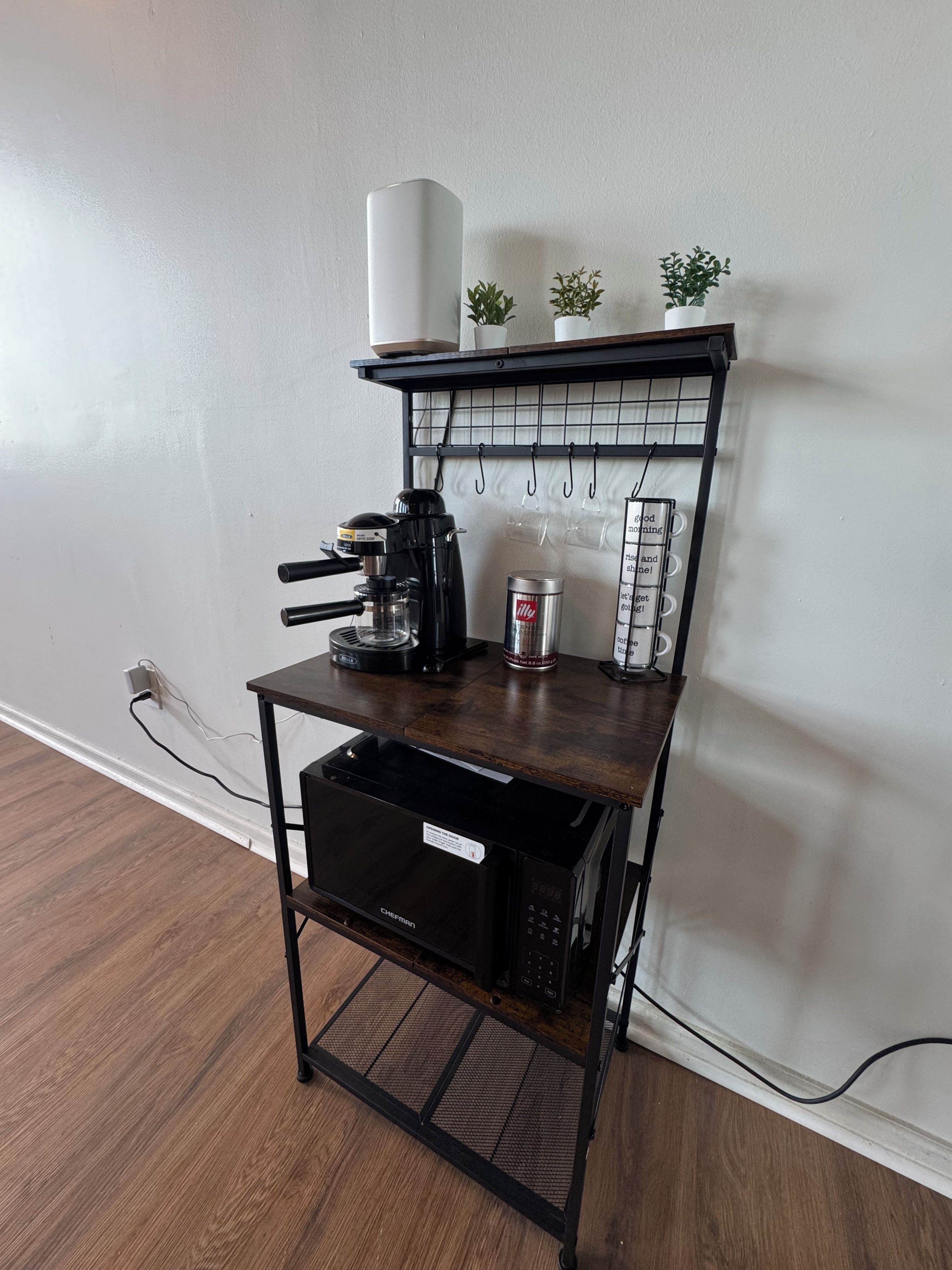 A kitchen area with a coffee maker, a can of coffee, a microwave, and a small shelf with plants.
