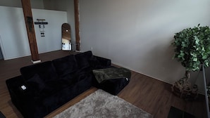 Ein Zimmer mit einem dunkelgrauen Sofa, einem grauen Teppich, Holzböden, einem Deckenventilator und einer Topfpflanze.