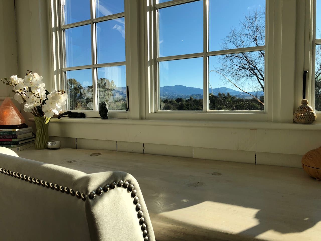 A room with a large window, a cushioned bench, a vase with flowers, and a small table with books and a lamp.