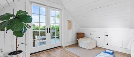 Una habitación con un ventanal grande, una planta en maceta, un pouf tejido y colchonetas de ejercicio.